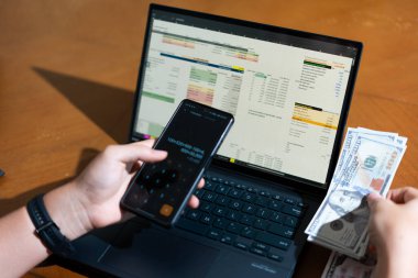 Bir kişi cep telefonu ve dizüstü bilgisayarı aynı anda kullanıyor. Cep telefonunda bir hesap makinesi var. Dizüstü bilgisayar ekranında çeşitli sayılar ve hesaplamalarla birlikte bir hesap tablosu gösteriliyor