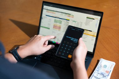Bir kişi cep telefonu ve dizüstü bilgisayarı aynı anda kullanıyor. Cep telefonunda bir hesap makinesi var. Dizüstü bilgisayar ekranında çeşitli sayılar ve hesaplamalarla birlikte bir hesap tablosu gösteriliyor