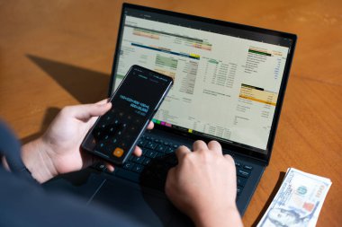 Bir kişi cep telefonu ve dizüstü bilgisayarı aynı anda kullanıyor. Cep telefonunda bir hesap makinesi var. Dizüstü bilgisayar ekranında çeşitli sayılar ve hesaplamalarla birlikte bir hesap tablosu gösteriliyor
