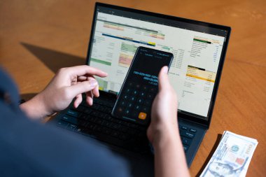 Bir kişi cep telefonu ve dizüstü bilgisayarı aynı anda kullanıyor. Cep telefonunda bir hesap makinesi var. Dizüstü bilgisayar ekranında çeşitli sayılar ve hesaplamalarla birlikte bir hesap tablosu gösteriliyor