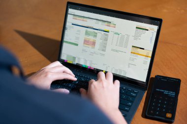 Bir kişi cep telefonu ve dizüstü bilgisayarı aynı anda kullanıyor. Cep telefonunda bir hesap makinesi var. Dizüstü bilgisayar ekranında çeşitli sayılar ve hesaplamalarla birlikte bir hesap tablosu gösteriliyor
