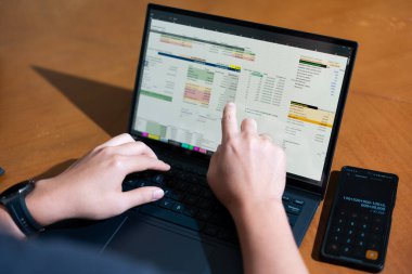 Bir kişi cep telefonu ve dizüstü bilgisayarı aynı anda kullanıyor. Cep telefonunda bir hesap makinesi var. Dizüstü bilgisayar ekranında çeşitli sayılar ve hesaplamalarla birlikte bir hesap tablosu gösteriliyor