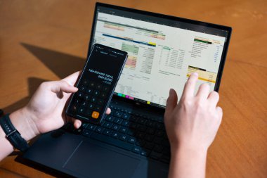 Bir kişi cep telefonu ve dizüstü bilgisayarı aynı anda kullanıyor. Cep telefonunda bir hesap makinesi var. Dizüstü bilgisayar ekranında çeşitli sayılar ve hesaplamalarla birlikte bir hesap tablosu gösteriliyor