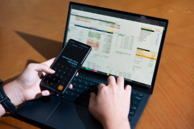 Bir kişi cep telefonu ve dizüstü bilgisayarı aynı anda kullanıyor. Cep telefonunda bir hesap makinesi var. Dizüstü bilgisayar ekranında çeşitli sayılar ve hesaplamalarla birlikte bir hesap tablosu gösteriliyor