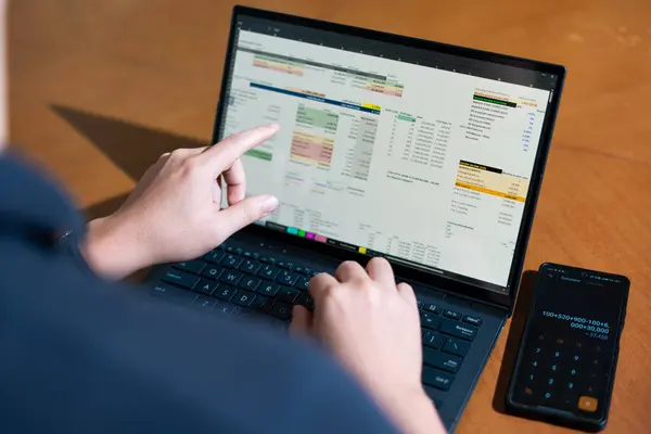 Bir kişi cep telefonu ve dizüstü bilgisayarı aynı anda kullanıyor. Cep telefonunda bir hesap makinesi var. Dizüstü bilgisayar ekranında çeşitli sayılar ve hesaplamalarla birlikte bir hesap tablosu gösteriliyor