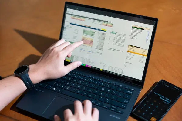 Bir kişi cep telefonu ve dizüstü bilgisayarı aynı anda kullanıyor. Cep telefonunda bir hesap makinesi var. Dizüstü bilgisayar ekranında çeşitli sayılar ve hesaplamalarla birlikte bir hesap tablosu gösteriliyor