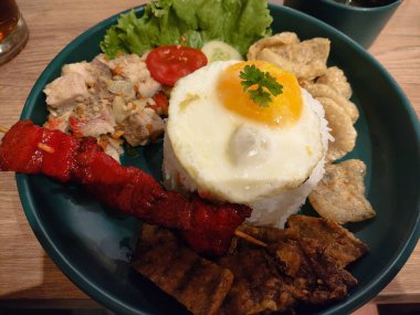 Bir tabak domuz yemeği ve pilavın üstünde sahanda yumurta. Tabak yeşil ve üzerinde çeşitli yiyecek maddeleri var.