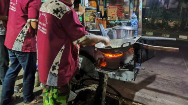 Bir adam tavada yemek pişirirken arkasında bir adam duruyor. Adam üzerinde 