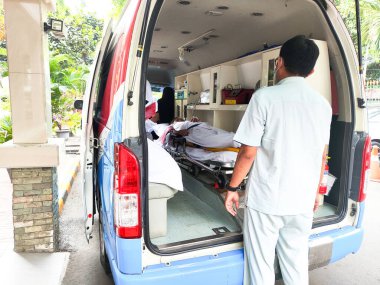 A man in a white shirt is standing in front of a white ambulance with a woman on a stretcher inside