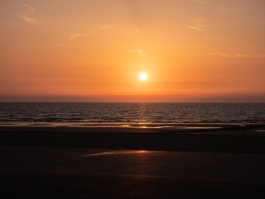 Kuzey Denizi kıyısında gün batımında dramatik bir gökyüzünün altında hafif dalgalar yuvarlanıyor. Turuncu ışık bulutlardan ve sudan yansıyarak barışçıl ve atmosferik bir deniz manzarası yaratır..