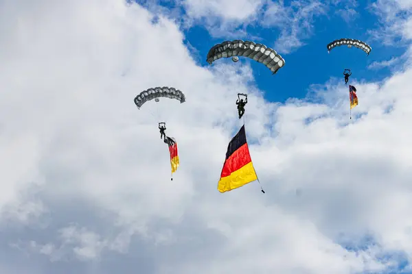 İki Alman Bundeswehr paraşüt birlikleri, açık mavi gökyüzüne karşı gururla Alman bayrağını taşıyarak, askeri hizmetteki gücü, birliği ve vatanseverliği simgeliyor..