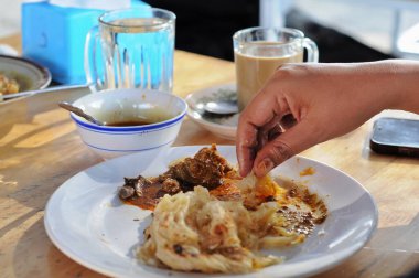 Bir adamın eli yemek için roti canai alıyor, ahşap masada.