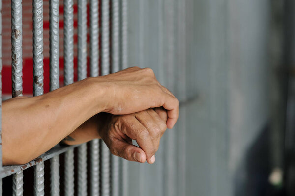  hands of a prisoner behind prison bars on black background