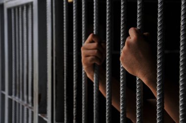 Mahkûmun elleri siyah arka planda parmaklıklar ardında.