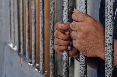 Mahkûmun elleri siyah arka planda parmaklıklar ardında.