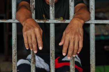 Mahkûmun elleri siyah arka planda parmaklıklar ardında.