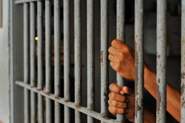 Mahkûmun elleri siyah arka planda parmaklıklar ardında.