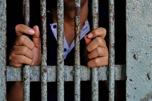 Mahkûmun elleri siyah arka planda parmaklıklar ardında.