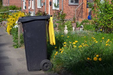Almanya 'da sarı papatyalı bir binaya karşı sarı kapaklı siyah bir çöp tenekesi.