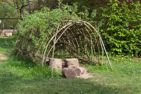 Dallardan ve yapraklardan yapılmış ev yapımı çadır. Ön planda yeşil çimlerin üzerinde kütükler var. Kuzey Almanya 'da.