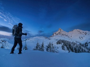 Night photo climber stands on top of a mountain in the snow and looks at the surrounding mountains with starry sky