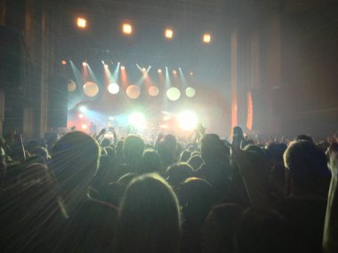 Konser çekimi. Işıklı bir sahnenin önünde büyük bir kalabalık bir grup performansını alkışlıyor. Bir grup insan sahnenin önünde duruyor.