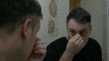 Genç bir adam banyoda aynanın önünde cımbızla kaşlarını yoluyor.