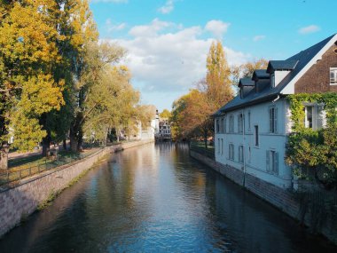 Strasbourg, Fransa. Manzaralı bir kanal, renkli binalarla dolu tarihi bir kasabanın içinden geçiyor. Yüzen evler kıyılar boyunca demirli, bazılarının pencere kutularında çiçekler var. Yeşil yapraklı ağaçlar kanal boyunca uzanır..