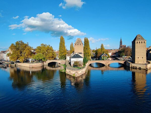 Ponts Couverts de Strasbourg. A stone bridge with three towers spans a blue river. Lush green trees line the riverbank on both sides.  In the background, a stone castle with a tall central tower sits on a hilltop.