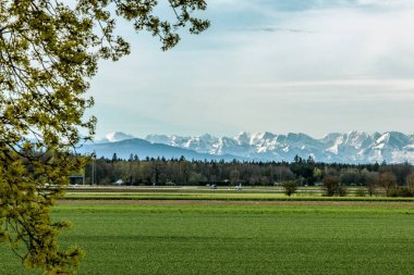 Münih 'in dışındaki Alp eteklerinin manzarası. Önde yeşil alanlar ve yapraksız ormanlar var. Dağlar odak noktası değil