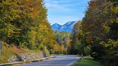 Bavyera dağlarının eteklerindeki dağların ve ormanların manzarası. Köy yolu ormandan geçiyor. Bir sonbahar akşamında ufuktaki dağlar. Mavi gökyüzü ve bulutlar.
