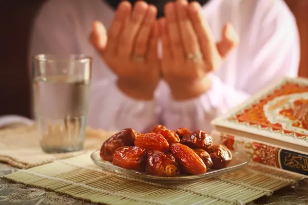 Allah 'a dua eden Müslüman kadınların elleriyle oruç yemeği ya da iftar tabağı. Masanın üzerinde bir bardak maden suyu olan tarihler. Geleneksel Ramazan, fast-break yemeği. Endonezya - 13 Mart 2024