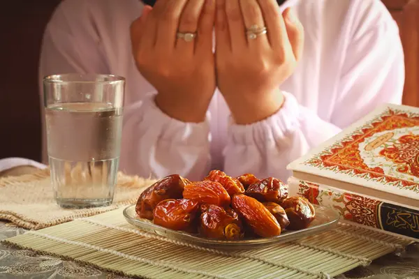 Allah 'a dua eden Müslüman kadınların elleriyle oruç yemeği ya da iftar tabağı. Masanın üzerinde bir bardak maden suyu olan tarihler. Geleneksel Ramazan, fast-break yemeği. Endonezya - 13 Mart 2024