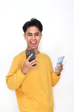 Genç Asyalı erkek gülümsüyor ve elinde cep telefonu ve beyaz arka planda izole edilmiş banknotlar tutuyor..