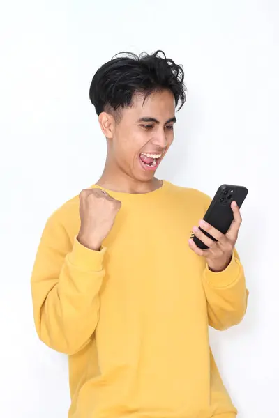 Cep telefonlu genç Asyalı adam zaferi beyaz arka planda yumrukla kutluyor.