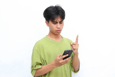 Yeşil tişörtlü genç adam gülümsüyor ve yazı yazarken telefonuna bakıyor, beyaz arka planda duruyor.