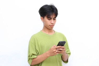 Yeşil tişörtlü genç adam gülümsüyor ve yazı yazarken telefonuna bakıyor, beyaz arka planda duruyor.