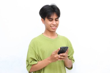 Yeşil tişörtlü genç adam gülümsüyor ve yazı yazarken telefonuna bakıyor, beyaz arka planda duruyor.