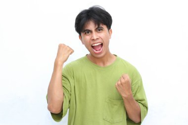 Yeşil gömlekli genç adam iki eli havada ve beyaz arka planda kararlı bir ifadeyle muzaffer bir jest yapıyor.