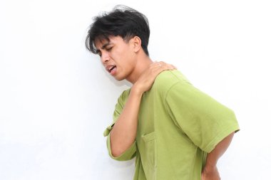 Yeşil gömlekli genç adam belini tutarken acı içinde kıvranıyor. Rahatsızlık ya da gerginlik yaşıyor gibi görünüyor. Beyaz arka plan ifadesini ve duruşunu vurguluyor.