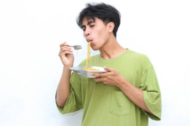 Yeşil tişörtlü genç adam beyaz arka planda çatallı bir tabak erişte yiyor. 