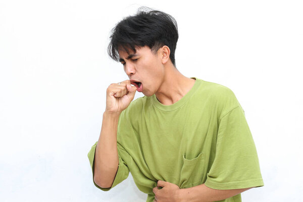 Young man in a green shirt coughs into his hand against a white background. He wears a smartwatch on his left wrist.