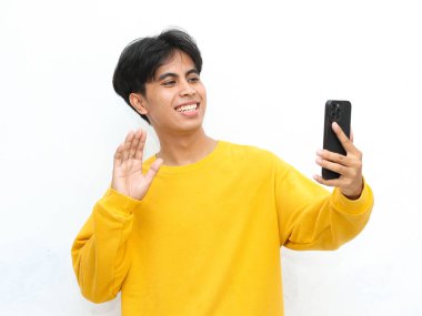 Sarı kazaklı genç adam beyaz arka planda akıllı telefonuyla selfie çekerken mutlu bir şekilde el sallıyor.