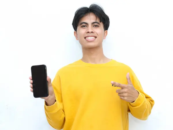 stock image Young man in a yellow sweater is holding a smartphone while pointing with the other hand while showing an expression of happiness on a white