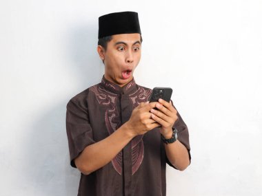 Beyaz bir arka planda izole edilmiş akıllı telefonu tutan ve işaret eden komik Asyalı Müslüman adam portresi. Ramazan konsepti.