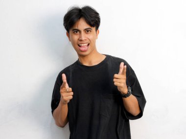 Siyah tişörtlü komik Asyalı genç adamın portresi beyaz arka planda izole edilmiş bir reklam için boş yeri işaret ediyor..