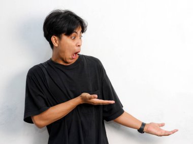 Siyah tişört giyen Asyalı genç bir adamın beyaz arka planda izole edilmiş bir şeye tepki göstermesinin komik bir portresi..