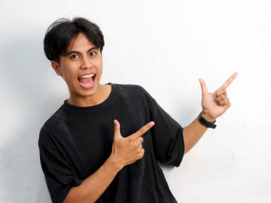 Siyah tişörtlü komik Asyalı genç adamın portresi beyaz arka planda izole edilmiş bir reklam için boş yeri işaret ediyor..