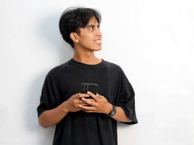 Siyah tişört giyen Asyalı genç bir adamın mutlu yüzü. Elinde akıllı telefon, boş alana bakıyor, beyaz arka planda izole edilmiş bir şeyi düşünüyor..