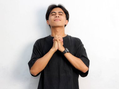 Siyah tişört giyen, beyaz arka planda elleri bağlı dua eden umut dolu Asyalı genç adamın portresi..  